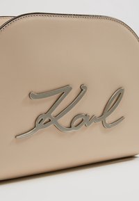 Beige handväska med rundad design, tillverkad av ett slätt material. Har en metallisk silverfärgad "Karl"-logotyp som är framträdande.