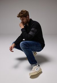 Svart kvartdraget tröja med texturerat tyg och en matt svart axelpatch, kombinerad med blå jeans och vita sneakers mot en ljus bakgrund.