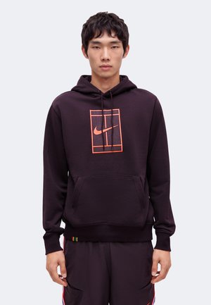 Giovane uomo che indossa una felpa nera con cappuccio con un logo arancione Nike all'interno di un grafico rettangolare, in piedi davanti a uno sfondo chiaro e uniforme.