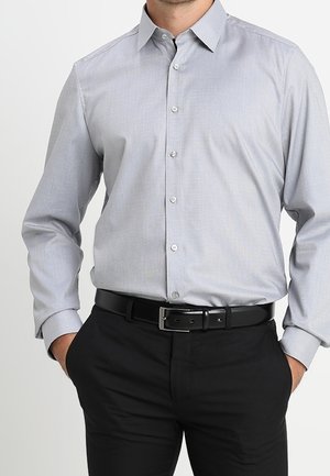 Light gray long-sleeve button-up shirt tucked into black pants with a black belt, hands resting in pants pockets.
