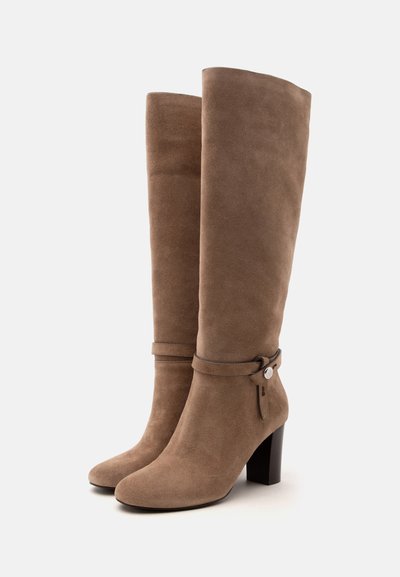 Bottes hautes en daim marron avec talons carrés et détails décoratifs de sangle et boucle près de la cheville, présentées sur fond blanc uni.