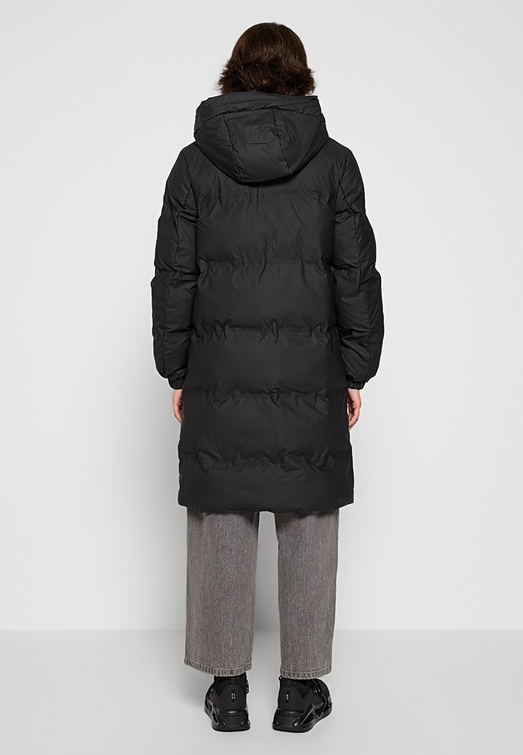 Manteau d'hiver noir long avec une capuche, présentant des panneaux cousus et des poignets élastiques. Porté par-dessus un pantalon large gris et des chaussures noires.