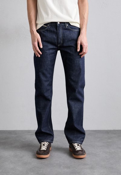 Dunkelblaue Jeans aus Denim mit geradem Bein, fünf Taschen und einem Knopfverschluss. Kombiniert mit braunen Wildlederschuhen und einem hellen Hemd.