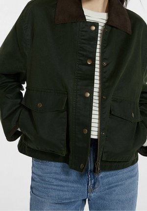 Personne portant une veste vert foncé avec un col marron, des boutons-pression en laiton, des poches à rabat, ouverte sur une chemise rayée blanche et noire et un jean bleu.