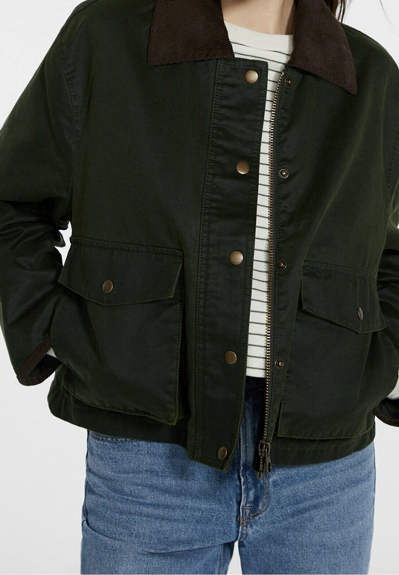 Personne portant une veste vert foncé avec un col marron, des boutons-pression en laiton, des poches à rabat, ouverte sur une chemise rayée blanche et noire et un jean bleu.