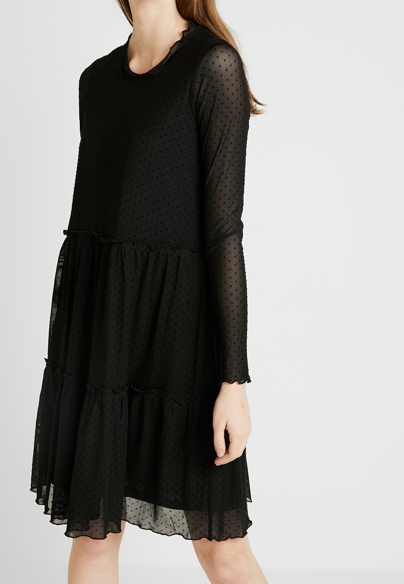 Femme portant une robe longue noire et transparente à manches longues avec de petits pois et un ourlet à volants, sur un fond clair uni.