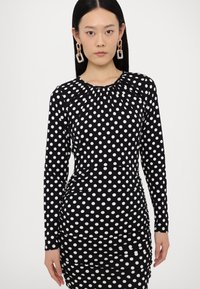 Black long-sleeve dress with white polka dots, gathered neckline, and ruched side detailing, featuring a fitted silhouette and smooth texture.
