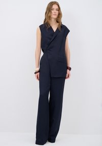 Mörkblå ärmlös jumpsuit med omslagdesign, har en framficka och vida byxor, tillverkad av ett texturerat tyg.