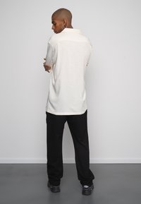 Camisa de manga larga color beige claro con cuello, textura sutil y dobladillo redondeado, combinada con pantalones negros y zapatos oscuros.