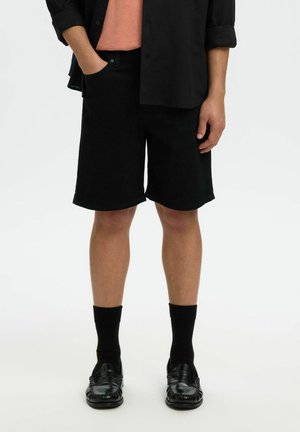 Person iført sorte shorts, sorte sokker, sorte loafers, en sort skjorte over en ferskenfarvet t-shirt, stående mod en hvid baggrund.