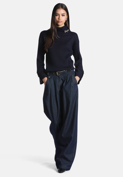 Femme portant un pull à col roulé côtelé noir avec une broche et un pantalon en jean sombre à jambes larges avec ceinture, mains dans les poches, debout sur un fond uni.