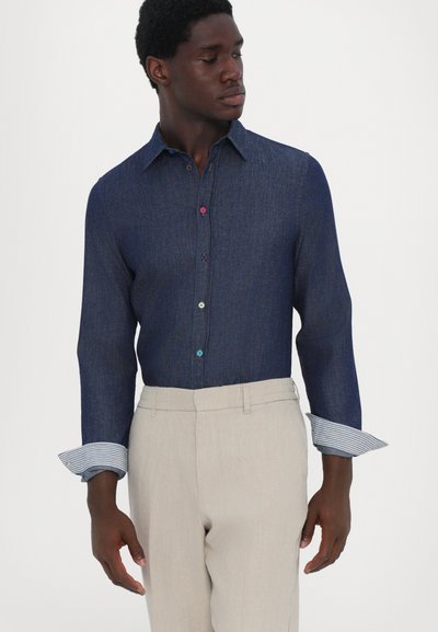 Navy blue denim shirt with a spread collar, featuring multicolored buttons. Stripe pattern on the inside of the rolled-up cuffs. Cream linen trousers.