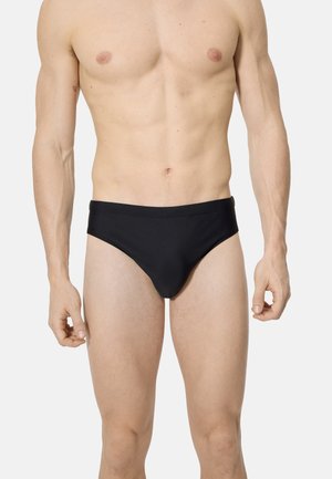 Torso masculino musculoso vistiendo bañadores negros lisos, de pie con los brazos relajados contra un fondo gris claro.