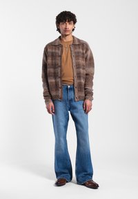 Brown plaid knitted cardigan over a brown shirt, paired with light blue flared jeans and brown leather shoes. Casual outfit, textured materials.