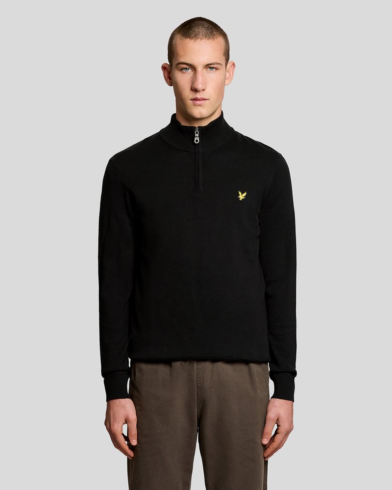 Νέος άνδρας με κοντά μαλλιά που φοράει μαύρο πουλόβερ με φερμουάρ στο λαιμό και μικρό κίτρινο λογότυπο πουλιού, καθώς και καφέ παντελόνι, στέκεται μπροστά σε γκρι φόντο.