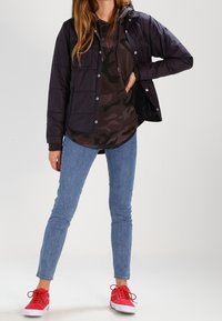 Chaqueta acolchada negra con botones a presión, llevada sobre una sudadera con capucha de camuflaje oscuro, combinada con jeans ajustados de mezclilla azul claros y zapatillas rojas brillantes.