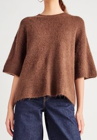 Pull marron ample en matière douce et duveteuse. Présente des manches courtes, un col rond et des finitions ourlet brut. Assorti à un jean bleu foncé.