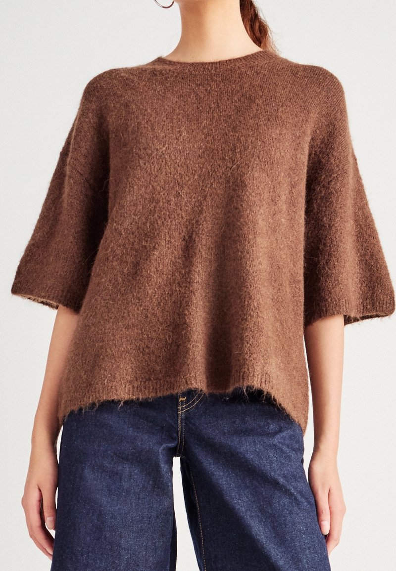 Pull marron ample en matière douce et duveteuse. Présente des manches courtes, un col rond et des finitions ourlet brut. Assorti à un jean bleu foncé.