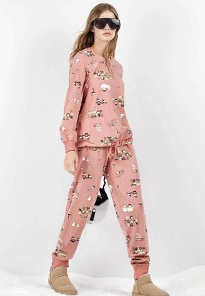 Femme portant un ensemble lounge rose imprimé de dessins animés, grandes lunettes de soleil noires, bottines beige à enfiler, debout sur fond blanc.