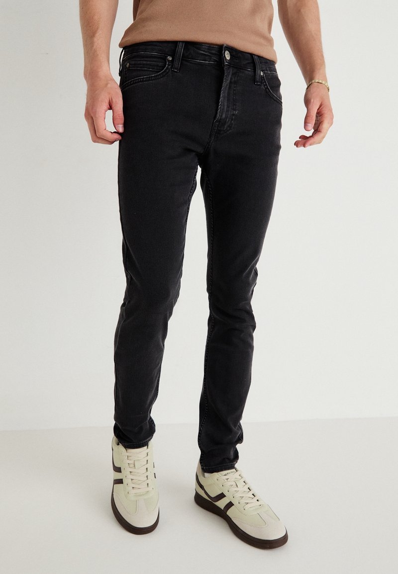 Mustat farkut, joissa on kapea malli, keskipitkä vyötärö ja kontrastiväriset tikkaus, yhdistettynä vaalean värisiin lenkkareihin.