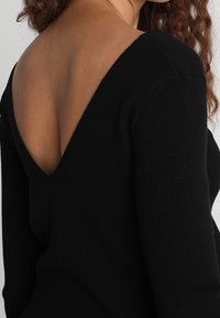 Svart ribbad tröja med djup V-rygg, långa ärmar och mjuk textur. Materialet verkar vara stickat med en åtsittande silhuett.
