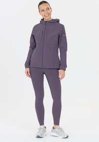 Vrouw die glimlacht, gekleed in een paarse hoodie, bijpassende leggings en zilveren hardloopschoenen, staande tegen een witte achtergrond.