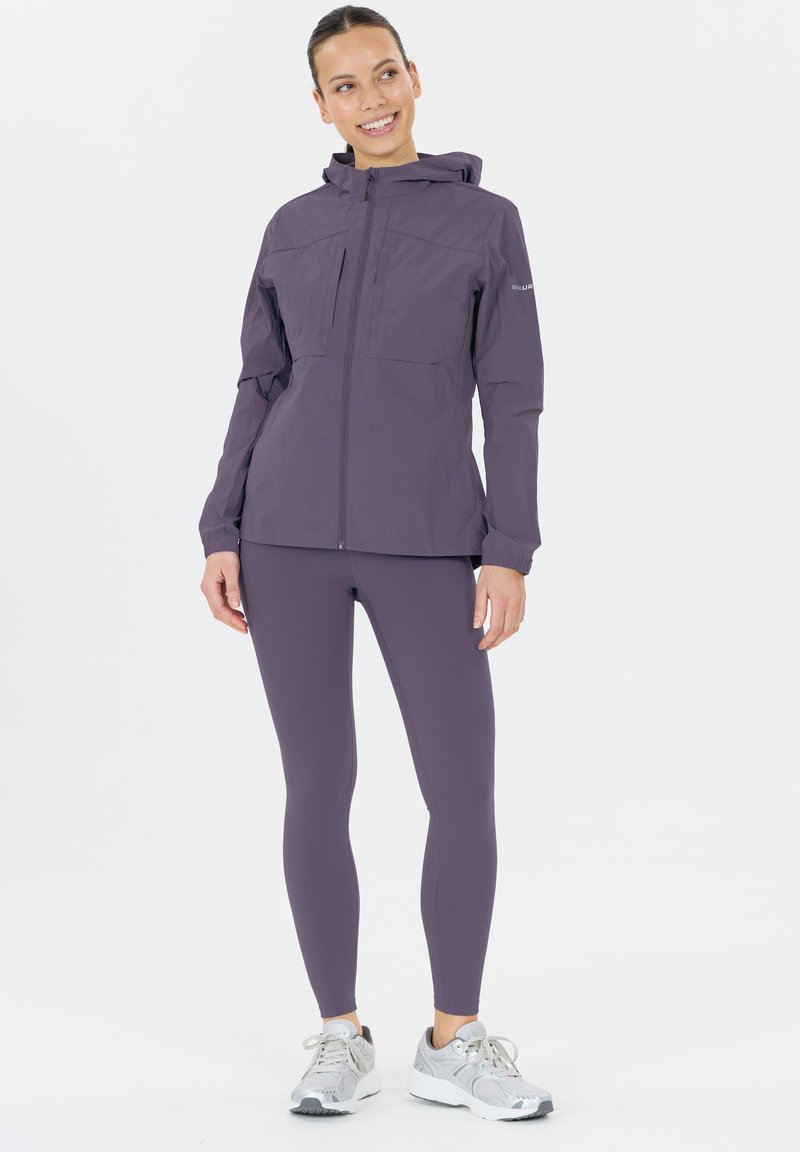 Vrouw die glimlacht, gekleed in een paarse hoodie, bijpassende leggings en zilveren hardloopschoenen, staande tegen een witte achtergrond.