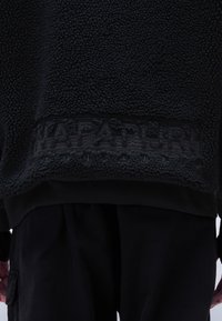 Sort fleece sweatervest med en tekstureret, blød overflade. Har et broderet logo ved kanten og sorte ribbede manchetter og talje.