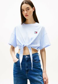Jasnoniebieska krótka koszulka z wiązaniem z przodu i krótkimi rękawami, z małą naszywką z logo. Połączona z ciemnoniebieskimi spodniami wysokiego stanu.
