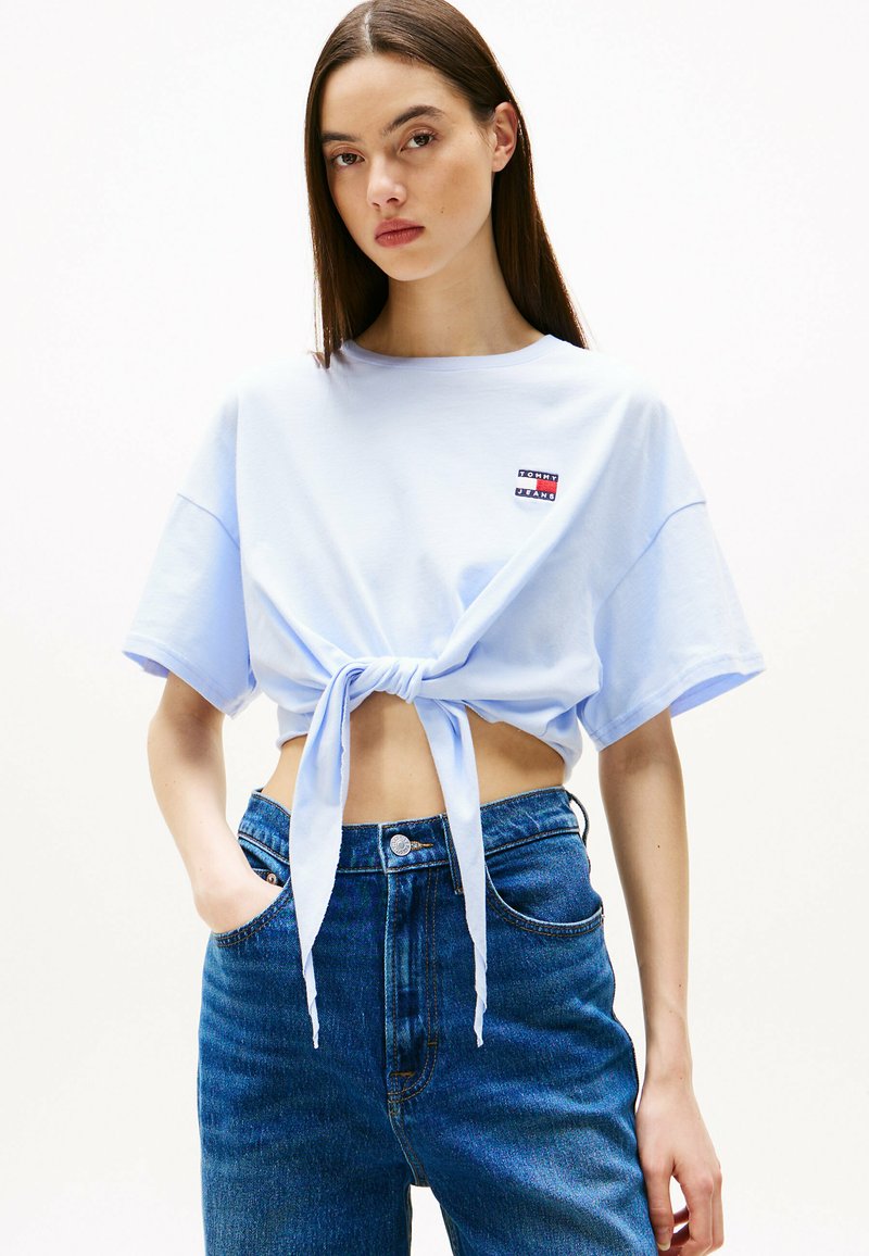 Jasnoniebieska krótka koszulka z wiązaniem z przodu i krótkimi rękawami, z małą naszywką z logo. Połączona z ciemnoniebieskimi spodniami wysokiego stanu.