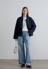 Μαύρο καπιτονέ bomber μπουφάν πάνω από λευκό t-shirt, συνδυασμένο με ανοιχτό μπλε τζιν με φαρδιά μπατζάκια και καφέ παπούτσια, κρατώντας μια μικρή μπεζ τσάντα.