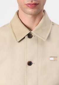 Beige cordfløyelsjakke med klassisk krage, to brune knapper og en liten logo-patch på brystet. Glatt, strukturert stoff.