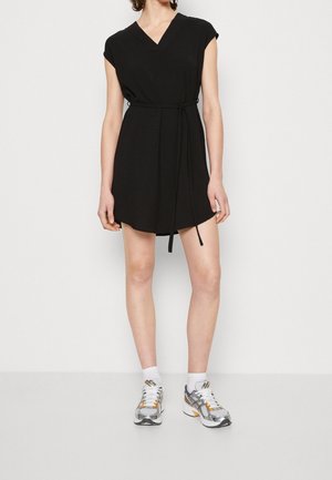 Femme portant une robe noire courte sans manches avec une ceinture à la taille, des chaussettes blanches et des chaussures de sport argentées et orange, debout sur un fond blanc.