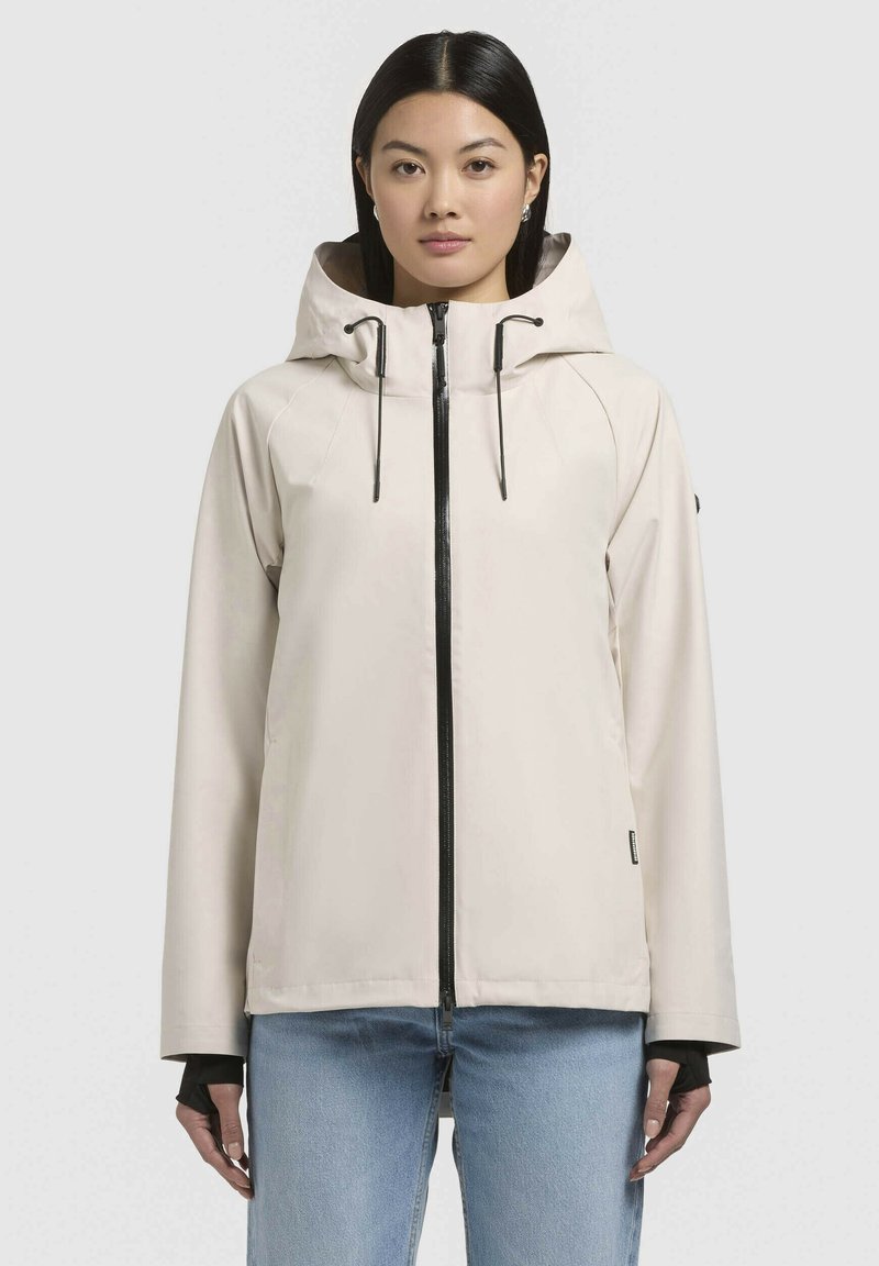Veste imperméable beige clair avec capuche, fermeture éclair noire et cordons ajustables. Dispose de manches longues et de trous pour les pouces pour un ajustement sécurisé.