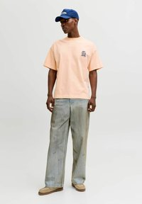 Bărbat purtând tricou grafic în culoarea piersicii, blugi largi, de culoare albastru deschis, cu pete de vopsea, pantofi bej și șapcă albastră, stând pe un fundal simplu.