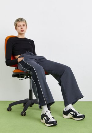 Persona con el pelo corto que lleva una camiseta corta negra y pantalones oscuros y holgados sentada relajada en una silla de oficina naranja sobre un suelo verde.