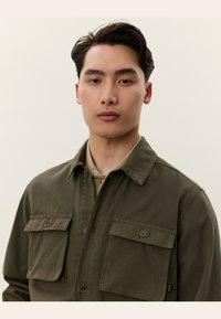 Giovane uomo con capelli scuri corti che indossa una giacca camicia verde oliva con bottoni su una camicia beige con colletto, sfondo neutro.