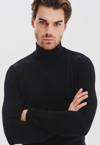 Czarny sweter z golfem, wykonany z gładkiego materiału dzianinowego, dopasowany krój, długie rękawy i ściągacz przy dekolcie, noszony z rękami skrzyżowanymi.