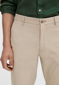 Mężczyzna w beżowych spodniach i zielonej koszuli z guzikami, z lewym ramieniem swobodnie opuszczonym przy boku.