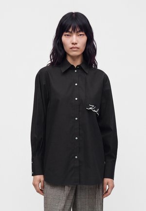Mulher de cabelo longo e escuro, vestindo uma camisa preta com botões e com um pequeno logótipo branco bordado "Karl", e calças aos quadrados.