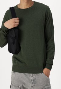 Maglione verde lavorato a maglia con polsini e orlo a coste, abbinato a una piccola borsa nera con zip appesa a una tracolla.
