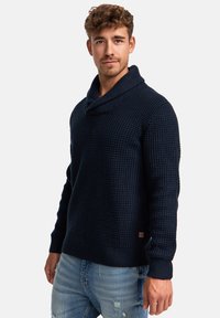 Granatowy sweter dzianinowy z kołnierzem szalowym, teksturowanym wzorem, ściągaczami przy rękawach i u dołu, z małą metką z logo po boku.