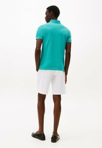 Türkises Poloshirt mit kurzen Ärmeln, strukturierter Stoff, kombiniert mit weißen Shorts. Schwarze Schuhe runden das Outfit ab. Rückansicht gezeigt.