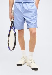 Άτομο που φοράει ανοιχτό μπλε αθλητικά σορτς και λευκά αθλητικά παπούτσια, κρατώντας μια ρακέτα τένις με μαύρο πλαίσιο και κίτρινα κορδόνια.