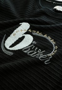 Schwarzer gerippter Stoff mit einer markanten weißen gestickten Zahl "6" und den Worten "by Ted Baker" in grau und goldenen Akzenten.