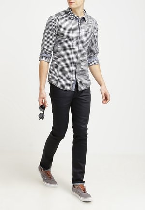 Man loopt, draagt een zwart-wit geruit overhemd met opgerolde mouwen, zwarte broek, grijze sneakers, houdt een zwarte zonnebril vast.