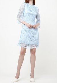 Ljusblå spetsklänning med cirkulära utskärningar, trekvarts ärmar med klockform och en scalloped nederkant, bärs med vita högklackade skor.