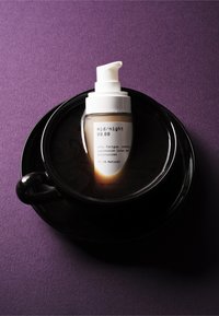 Gjennomsiktig glassflaske med pumpe, hvit pumpe og etikett, inneholdende lys krem, plassert i en svart kopp på lilla bakgrunn.