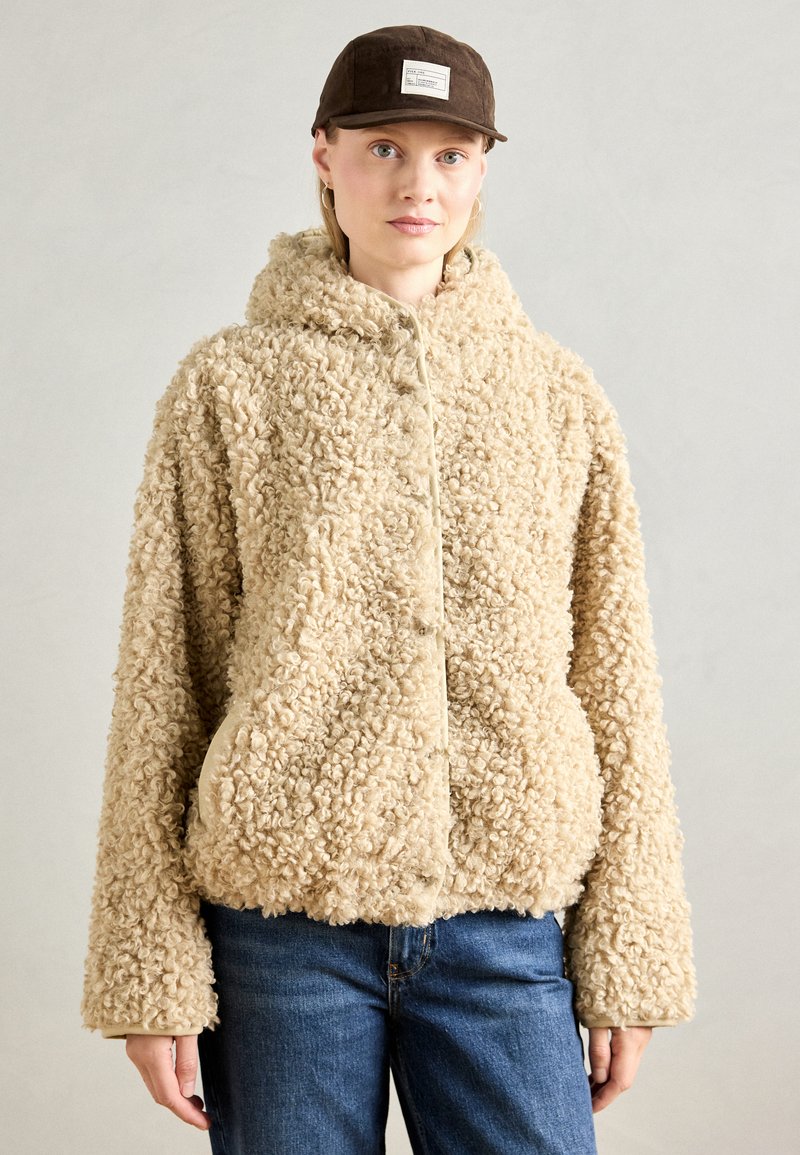 Kvinne i en beige krøllete fleecejakke med hette, blå jeans og en brun caps med en hvit lapp foran.