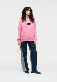 Ροζ φαρδιά μπλούζα με κουκούλα και μπροστινή τσέπη, με μαύρο λογότυπο, σε συνδυασμό με τζιν παντελόνι φαρδιάς γραμμής με αντίθετες πλαϊνές λεπτομέρειες και μαύρα παπούτσια.